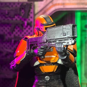 May include: A science fiction action figure wearing bright orange and black armor with a matching helmet. The figure holds a large, metallic silver blaster rifle with both hands. The armor features a chest plate, shoulder guards, and a belt.