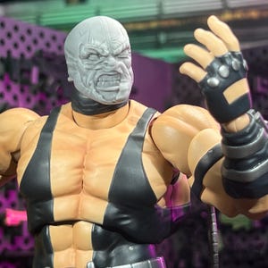 May include: A detailed action figure of a muscular character with a gray mask and black and gray attire. The figure has a stern expression and is posed with one arm raised, showcasing a black glove with silver studs. The figure's skin tone is tan.