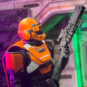 Op de afbeelding: Een plastic actiefiguur met een feloranje helm en bijpassend borstpantser over een zwart pak. Het figuur houdt een groot, gedetailleerd zwart geweer in de hand. De helm is voorzien van een donker vizier en het pantser heeft grijze accenten.