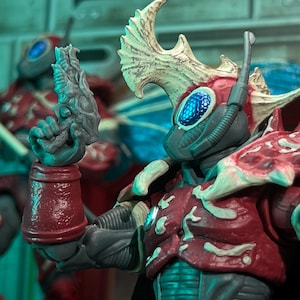 Peut inclure: Gros plan sur une figurine rouge et grise avec des détails complexes. La figurine porte un casque avec un œil bleu et tient une arme. Fait partie d'une collection de figurines.