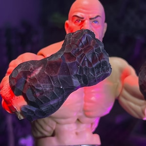 May include: A muscular action figure with a bald head and light skin tone, featuring a large, dark, textured fist with a faceted, rock-like surface. The figure is illuminated by vibrant red and purple lighting, highlighting its defined chest muscles.