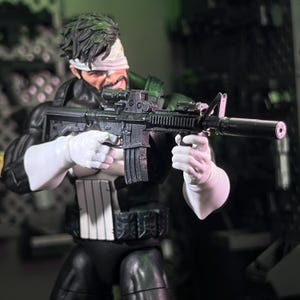 May include: A male action figure with dark hair and a white bandage over one eye, wearing a black suit with a white skull emblem on the chest. The figure holds a black assault rifle with a scope and suppressor, featuring white gloves and a utility belt.