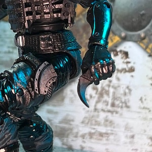 May include: A detailed action figure with a metallic teal and black color scheme. The figure is holding a curved blade. The figure has intricate armor and a utility belt. The overall theme is science fiction or fantasy.
