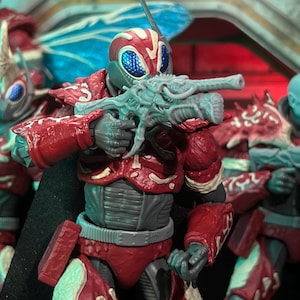 Peut inclure: Figurine d'action rouge et grise avec un casque et une armure détaillés en forme d'insecte, tenant une arme bleu clair. La figurine a une cape noire et fait partie d'un groupe de figurines similaires, suggérant un thème de science-fiction ou de fantaisie.