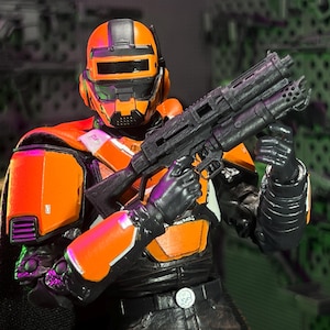 Op de afbeelding: Een beweegbaar actiefiguur met een feloranje en zwarte tactische helm en bijpassende bepantsering over een zwart pak. Het figuur houdt een groot zwart geweer vast en heeft een riem met een gesp voorzien van een schedel-embleem.
