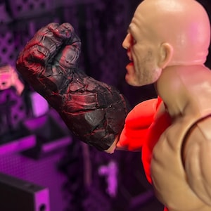 Könnte beinhalten: Eine Kunststoff-Actionfigur mit Glatze und gebräunter Haut, die einen großen, überdimensionalen rechten Arm mit einer dunklen, rissigen, felsartigen Textur aufweist. Die Figur ist im Profil dargestellt, mit erhobener, rot beleuchteter Faust.