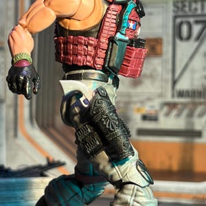 Könnte beinhalten: Detaillierte Actionfigur eines Soldaten mit roter taktischer Weste, schwarzen Handschuhen und verzierter Beinrüstung. Die Figur hat einen muskulösen Körperbau und ist dynamisch posiert. Das Design umfasst einen blau-roten Aufnäher auf der Weste.