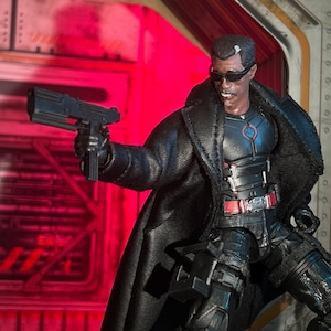 May include: A detailed action figure of a character in a black coat, sunglasses, and tactical gear, holding a black submachine gun. The figure has a determined expression, set against a red-lit background.