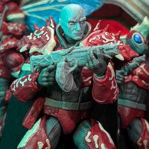 Peut inclure: Figurine d'action détaillée d'un humanoïde à la peau bleue tenant une arme futuriste. La figurine porte une armure rouge et blanche avec des motifs complexes. Le personnage a une expression sévère, sur fond flou.