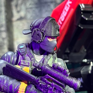 Puede incluir: Figura de acción negra de un soldado que lleva una gorra negra, una máscara y equipo táctico. La figura sostiene un rifle negro con mira telescópica. El fondo incluye un objeto rojo con texto blanco.