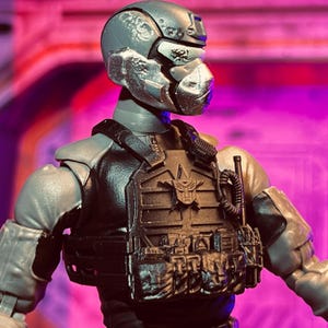 May include: A gray action figure of a soldier wearing a helmet and tactical vest. The helmet has a silver face guard. The vest has pouches and a symbol on the front. The figure is posed against a pink and purple background.