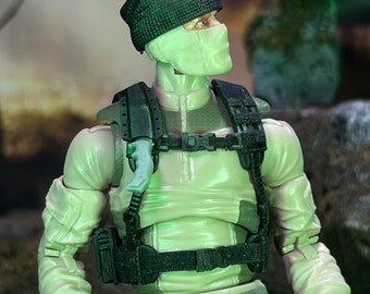 May include: A detailed action figure of a soldier in a light green uniform, black tactical vest, and a black beanie. The figure has a white face covering and a knife attached to the vest. The figure is posed in a dynamic stance.