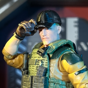 May include: A detailed action figure of a soldier wearing a black cap, tan and green tactical vest, and gloves. The figure is holding the cap with a gloved hand. The figure has a serious expression.