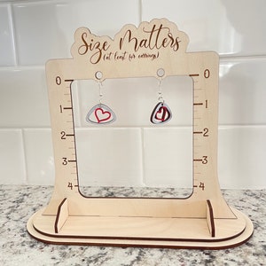May include: A light-colored wooden earring display stand with the text Size Matters (at least for earrings) and a 0 to 4 inch scale on both sides. Two dangling earrings with silver hooks and red and grey heart-shaped charms hang from the top.