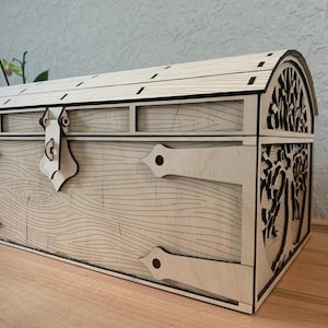 Puede incluir: Un cofre rectangular de madera con tapa curva, acabado en tono bronceado claro y patrones de veta de madera cortados con láser. El frente incluye un pestillo central y detalles de bisagras decorativas, con un diseño de árbol calado en el lateral.