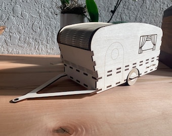 May include: A light-colored wooden model of a travel trailer featuring a hitch, a single wheel, and engraved details including a window with curtains and a small door. The structure is assembled with visible interlocking joints and dark edges.