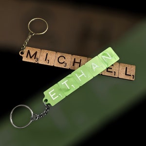 May include: Two keychains featuring letter tiles. One keychain is made of brown cork-like material spelling the name MICHAEL with black letters and numbers. The other is light green plastic spelling ETHAN with white letters and numbers.
