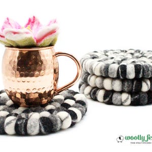 Puede incluir: Una taza de Moscow Mule de cobre con una flor de fieltro y una pila de posavasos de bolas de fieltro negras, blancas y grises. Los posavasos tienen aproximadamente 10 cm de diámetro. La imagen también incluye el logotipo "woolly felt".