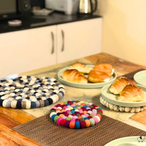 May include: Two round trivets made of felted wool balls. One trivet features a gradient of blue, gray, and white tones, while the smaller one displays a vibrant multicolored pattern. Both items are placed on a wooden surface near plates of food.