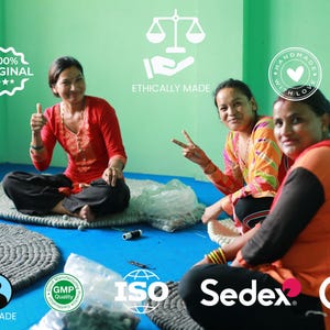 Puede incluir: Tres mujeres sentadas en el suelo, sonriendo, rodeadas de alfombras circulares grises hechas a mano. La imagen incluye texto que dice "100% Original", "Ethically Made", "Handmade with Love", "Fairtrade", "GMP Quality", "ISO", "Sedex" y "CE".