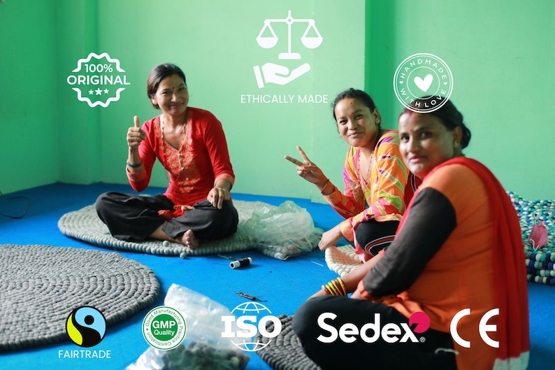 Puede incluir: Tres mujeres sentadas en el suelo, sonriendo, rodeadas de alfombras circulares grises hechas a mano. La imagen incluye texto que dice "100% Original", "Ethically Made", "Handmade with Love", "Fairtrade", "GMP Quality", "ISO", "Sedex" y "CE".
