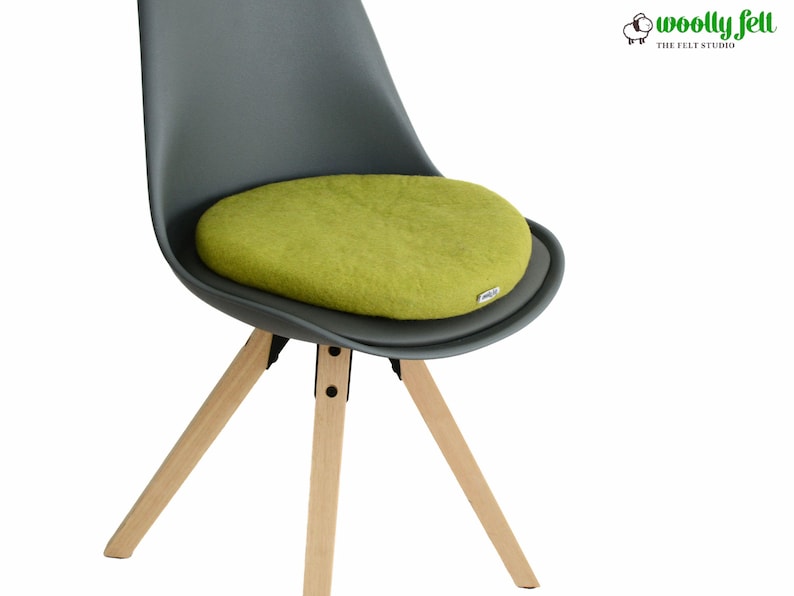 K&ouml;nnte beinhalten: Ein rundes, lindgr&uuml;nes Filz-Sitzkissen auf einem dunkelgrauen geformten Stuhl mit hellen Holzbeinen. Das Kissen hat ein kleines wei&szlig;es Etikett an der Seite. Das Logo von Woolly Felt The Felt Studio ist in der oberen rechten Ecke zu sehen.