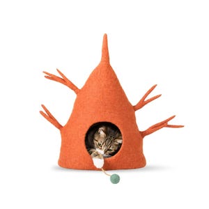 May include: A cone-shaped orange felt cat cave with protruding branch-like structures on the sides. A tabby cat is inside the circular opening, reaching out with a paw to touch a small green ball attached to a string. The text woolly felt is visible.