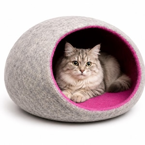 Pode incluir: Uma cama para gatos em forma de cúpula feita de feltro cinza claro com um interior magenta vibrante. Um gato de pelo comprido, cor tigrado prateado e olhos verdes, descansa dentro da abertura circular desta toca macia e arredondada.