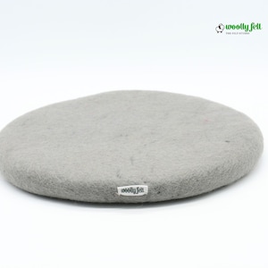 K&ouml;nnte beinhalten: Ein kreisf&ouml;rmiges, hellgraues Filzpolster mit strukturierter Oberfl&auml;che. Am Rand befindet sich ein kleines wei&szlig;es Stoffetikett mit der schwarzen Aufschrift "woolly felt". Das Markenlogo "woolly felt" mit einem Schafsymbol ist oben zu sehen.