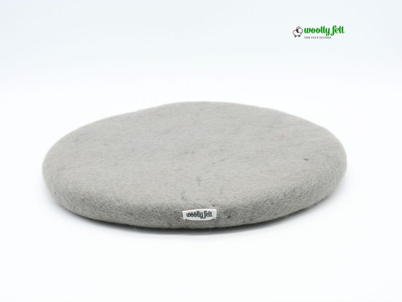 K&ouml;nnte beinhalten: Ein kreisf&ouml;rmiges, hellgraues Filzpolster mit strukturierter Oberfl&auml;che. Am Rand befindet sich ein kleines wei&szlig;es Stoffetikett mit der schwarzen Aufschrift "woolly felt". Das Markenlogo "woolly felt" mit einem Schafsymbol ist oben zu sehen.