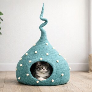 May include: A teal felted cat cave with a tall, spiralling top and small white pom-poms scattered across the exterior. The structure has a circular opening at the front, revealing a fluffy grey and white tabby cat resting inside the cosy enclosure.