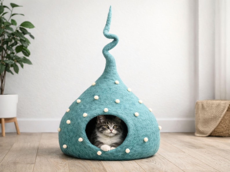 May include: A teal felted cat cave with a tall, spiralling top and small white pom-poms scattered across the exterior. The structure has a circular opening at the front, revealing a fluffy grey and white tabby cat resting inside the cosy enclosure.