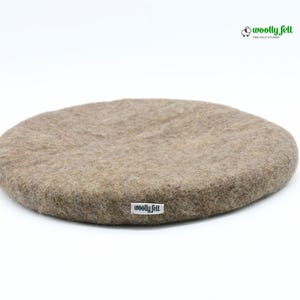 K&ouml;nnte beinhalten: Ein rundes, beigefarbenes Filzkissen mit strukturierter Oberfl&auml;che. An der Seite ist ein kleines wei&szlig;es Stoffetikett mit dem schwarzen Schriftzug "woolly felt" angebracht. In der oberen rechten Ecke befindet sich ein Logo mit einem Schafsymbol und dem Text "woolly felt".