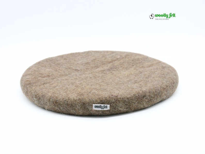 K&ouml;nnte beinhalten: Ein rundes, beigefarbenes Filzkissen mit strukturierter Oberfl&auml;che. An der Seite ist ein kleines wei&szlig;es Stoffetikett mit dem schwarzen Schriftzug "woolly felt" angebracht. In der oberen rechten Ecke befindet sich ein Logo mit einem Schafsymbol und dem Text "woolly felt".