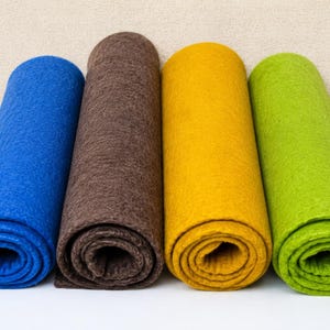 May include: Four rolls of felt in blue, brown, yellow, and green. The felt is a thick, textured material, each roll neatly coiled. The rolls are arranged side by side against a neutral background. The felt is likely measured in centimetres.