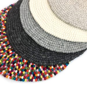 May include: A collection of felted wool trivets in various colors. The trivets are round and made of small, tightly packed wool balls. Colors include a vibrant multicolored option, dark gray, light gray, off-white, and a lighter gray.