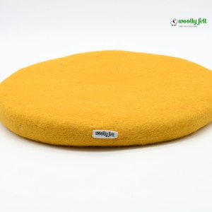 K&ouml;nnte beinhalten: Ein kreisf&ouml;rmiges, senfgelbes Filzpolster mit strukturierter Oberfl&auml;che. Am Rand ist ein kleines wei&szlig;es Stoffetikett mit dem schwarzen Schriftzug "woolly felt" angen&auml;ht. Oben rechts befindet sich ein Logo mit Schafsymbol und "woolly felt".