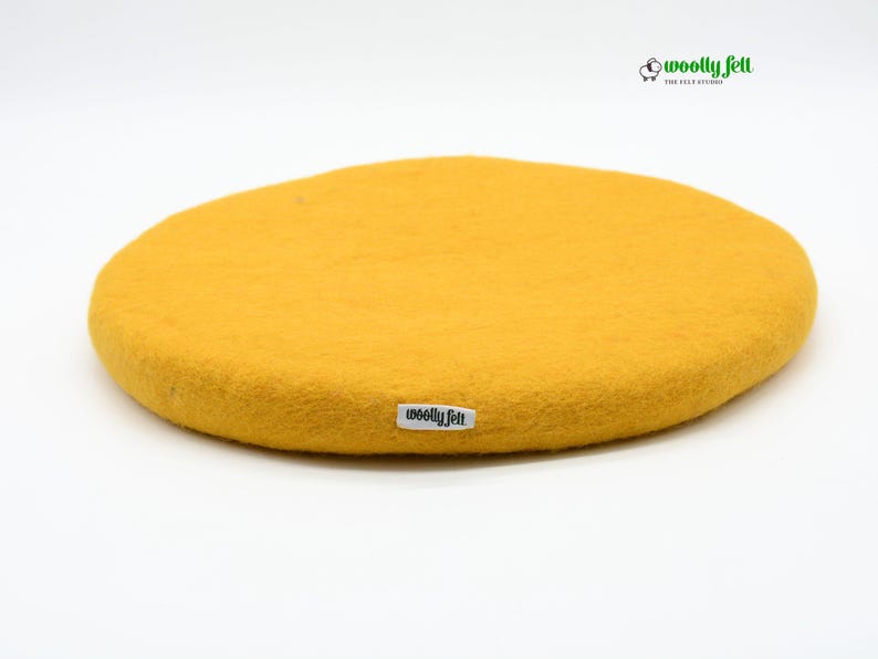 K&ouml;nnte beinhalten: Ein kreisf&ouml;rmiges, senfgelbes Filzpolster mit strukturierter Oberfl&auml;che. Am Rand ist ein kleines wei&szlig;es Stoffetikett mit dem schwarzen Schriftzug "woolly felt" angen&auml;ht. Oben rechts befindet sich ein Logo mit Schafsymbol und "woolly felt".