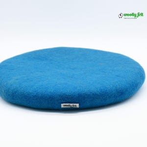 K&ouml;nnte beinhalten: Ein rundes, leuchtend blaues Kissen aus gefilzter Wolle mit einer weichen, strukturierten Oberfl&auml;che. Ein kleines wei&szlig;es Stoffetikett mit dem schwarzen Text 'woolly felt' ist an der Seite angebracht. Oben rechts befindet sich ein Logo.