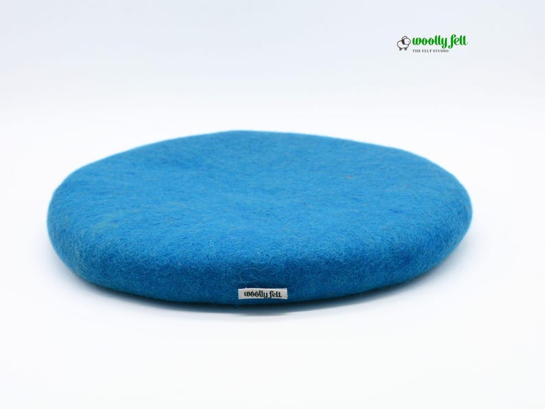 K&ouml;nnte beinhalten: Ein rundes, leuchtend blaues Kissen aus gefilzter Wolle mit einer weichen, strukturierten Oberfl&auml;che. Ein kleines wei&szlig;es Stoffetikett mit dem schwarzen Text 'woolly felt' ist an der Seite angebracht. Oben rechts befindet sich ein Logo.