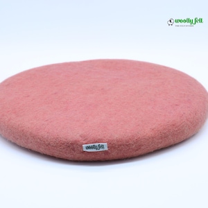 K&ouml;nnte beinhalten: Ein rundes, altrosa Sitzkissen aus Filz mit einer strukturierten, matten Oberfl&auml;che. Ein kleines wei&szlig;es Stoffetikett mit dem schwarzen Text "woolly felt" ist an der Seite angebracht. In der oberen rechten Ecke befindet sich ein Logo mit einem Schaf und dem Text "woolly felt".