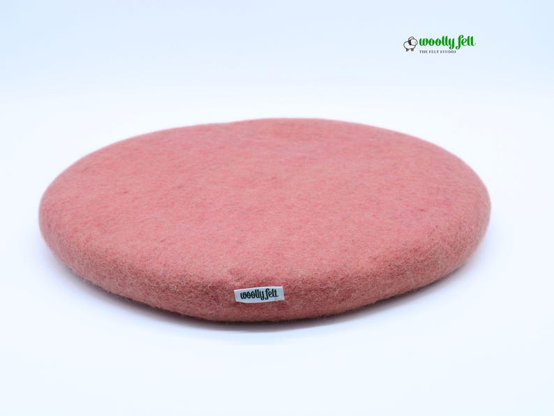K&ouml;nnte beinhalten: Ein rundes, altrosa Sitzkissen aus Filz mit einer strukturierten, matten Oberfl&auml;che. Ein kleines wei&szlig;es Stoffetikett mit dem schwarzen Text "woolly felt" ist an der Seite angebracht. In der oberen rechten Ecke befindet sich ein Logo mit einem Schaf und dem Text "woolly felt".