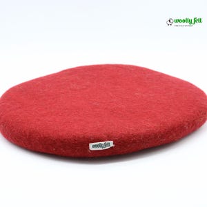 K&ouml;nnte beinhalten: Ein rundes, rotes Filzpolster aus Wolle mit strukturierter Oberfl&auml;che. An der Seite befindet sich ein kleines wei&szlig;es Stoffetikett mit dem schwarzen Text "woolly felt". In der oberen rechten Ecke ist ein Logo mit Schafsymbol und "woolly felt THE FELT STUDIO".