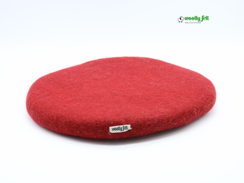 K&ouml;nnte beinhalten: Ein rundes, rotes Filzpolster aus Wolle mit strukturierter Oberfl&auml;che. An der Seite befindet sich ein kleines wei&szlig;es Stoffetikett mit dem schwarzen Text "woolly felt". In der oberen rechten Ecke ist ein Logo mit Schafsymbol und "woolly felt THE FELT STUDIO".