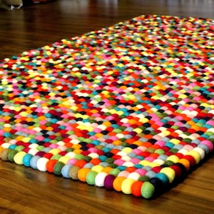 May include: A colorful rug made of felt balls in various shades of red, orange, yellow, green, blue, purple, pink, and white. The balls are arranged in a grid pattern, creating a visually appealing and textured surface.