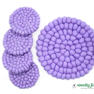May include: A collection of lavender felt coasters. The coasters are round with a textured, bubbly surface. Four coasters are stacked, and one is separate. The image is on a white background. The brand logo "woolly felt" is in the bottom right corner.