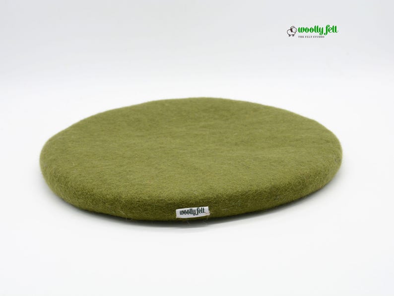 K&ouml;nnte beinhalten: Ein rundes, olivgr&uuml;nes Kissen aus gefilzter Wolle mit einer weichen, strukturierten Oberfl&auml;che. Ein kleines wei&szlig;es Stoffetikett mit dem schwarzen Text "woolly felt" ist am Rand befestigt. Das Logo "woolly felt THE FELT STUDIO" mit einem Schaf-Symbol ist dar&uuml;ber zu sehen.