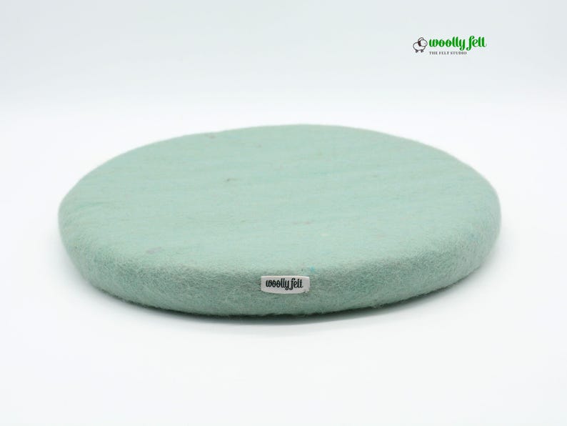 K&ouml;nnte beinhalten: Ein rundes, mintgr&uuml;nes Filzkissen aus Wolle mit einer dichten, strukturierten Oberfl&auml;che. Ein kleines wei&szlig;es Stoffetikett mit dem schwarzen Text "woolly felt" ist an der Seite angebracht. Oben rechts befindet sich ein Logo mit Schafsymbol.