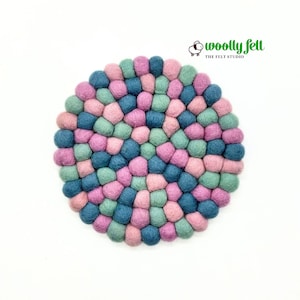 May include: A circular trivet made of numerous small, round felt balls in shades of pink, teal, and dark blue. The text "woolly felt THE FELT STUDIO" with a small sheep icon is printed in green and black above the circular felt ball arrangement.