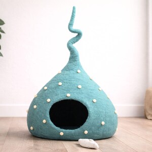 May include: A teal felted pet cave with a tall, spiralling top and a wide, circular opening. The exterior is decorated with small, cream-coloured felted dots scattered across the surface. A small, light-coloured felted toy lies on the floor.
